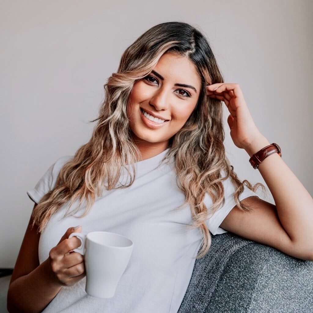 Marianna Teudor A smiling woman with wavy blonde hair, wearing a white t-shirt, sitting on a gray couch against a plain background.