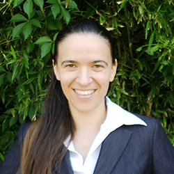 Melissa A smiling woman with long dark hair wearing a dark blazer and white shirt, standing in front of green leafy plants.